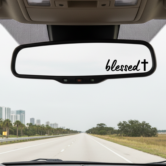 Car interior with a 'blessed' decal on the rearview mirror, driving on a road with trees and buildings in the background.