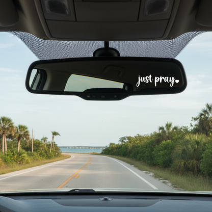Car interior with 'just pray' sticker on the rearview mirror, road ahead with greenery.