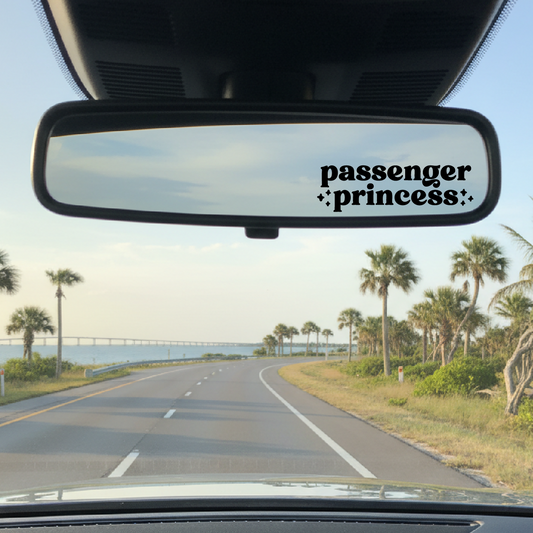Car interior with 'passenger princess' decal on rearview mirror, palm trees and road visible outside.