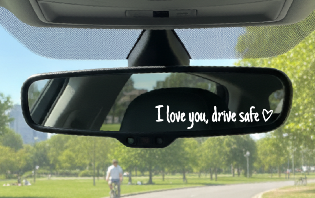 Car rearview mirror with 'I love you, drive safe' sticker in a park setting
